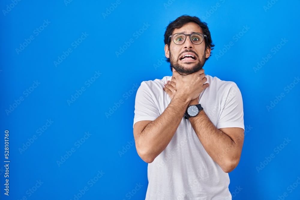 Handsome latin man standing over blue background shouting and suffocate because painful strangle. health problem. asphyxiate and suicide concept.