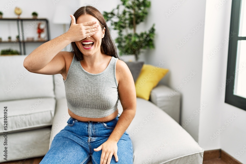 Young brunette woman sitting on the sofa at home smiling and laughing with hand on face covering eyes for surprise. blind concept. Young brunette woman sitting on the sofa at home smiling and laughing with hand on face covering eyes for surprise. blind concept.