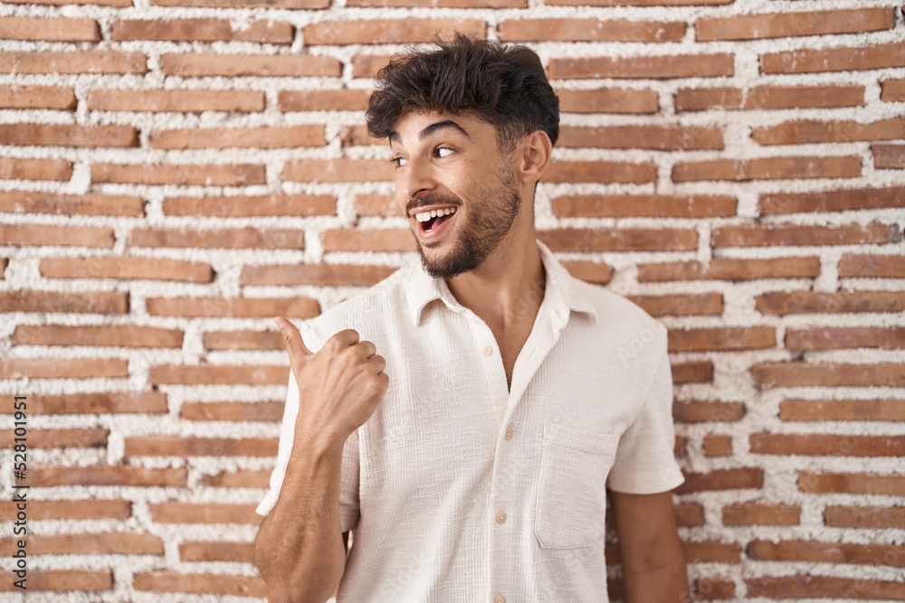 Arab man with beard standing over bricks wall background smiling with happy face looking and pointing to the side with thumb up. Arab man with beard standing over bricks wall background smiling with happy face looking and pointing to the side with thumb up.