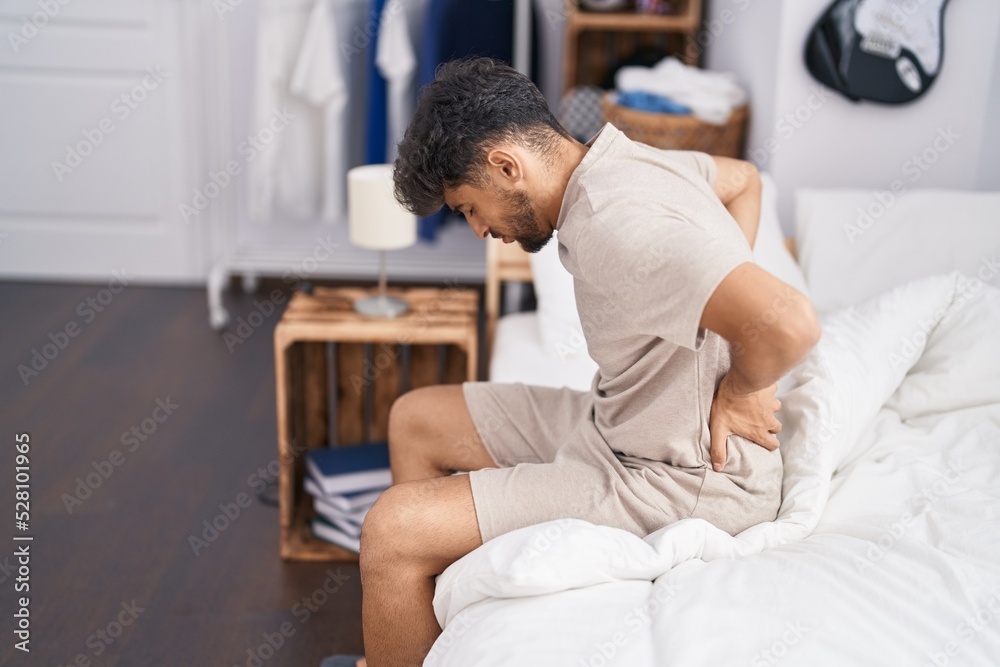 Young arab man suffering for backache sitting on bed at bedroom