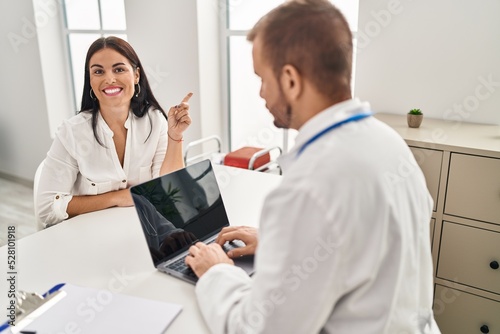 Young hispanic woman at the doctor smiling happy pointing with hand and finger to the side