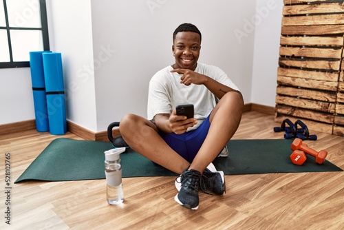 Young african man sitting on training mat at the gym using smartphone pointing to the back behind with hand and thumbs up  smiling confident