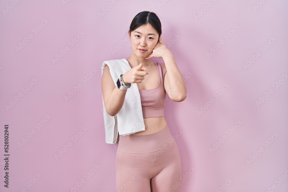 Chinese young woman wearing sportswear and towel smiling doing talking on the telephone gesture and pointing to you. call me.