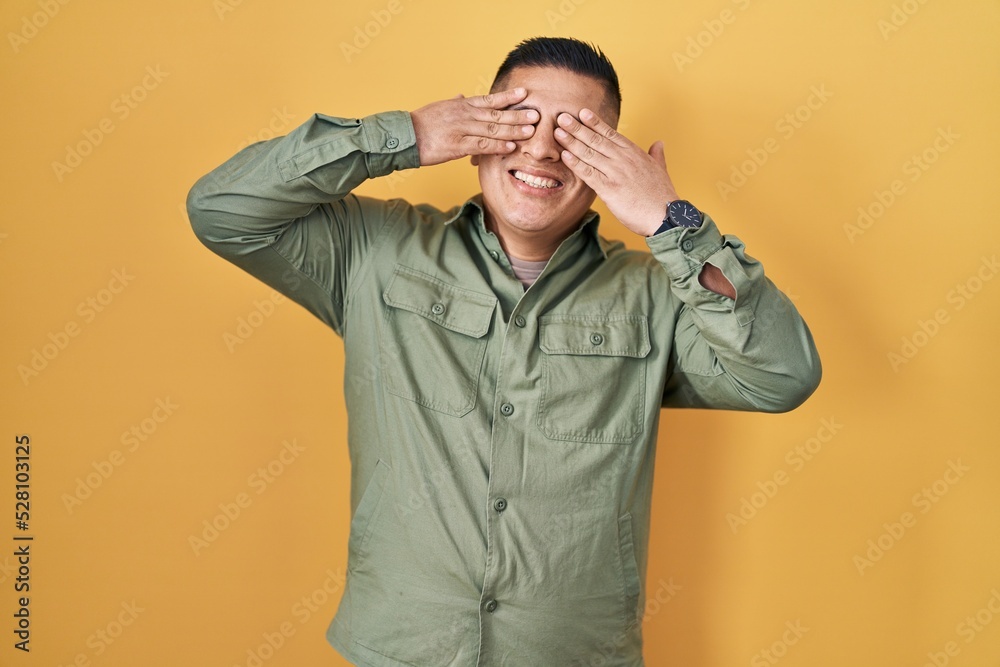 Hispanic young man standing over yellow background covering eyes with hands smiling cheerful and funny. blind concept. Hispanic young man standing over yellow background covering eyes with hands smiling cheerful and funny. blind concept.