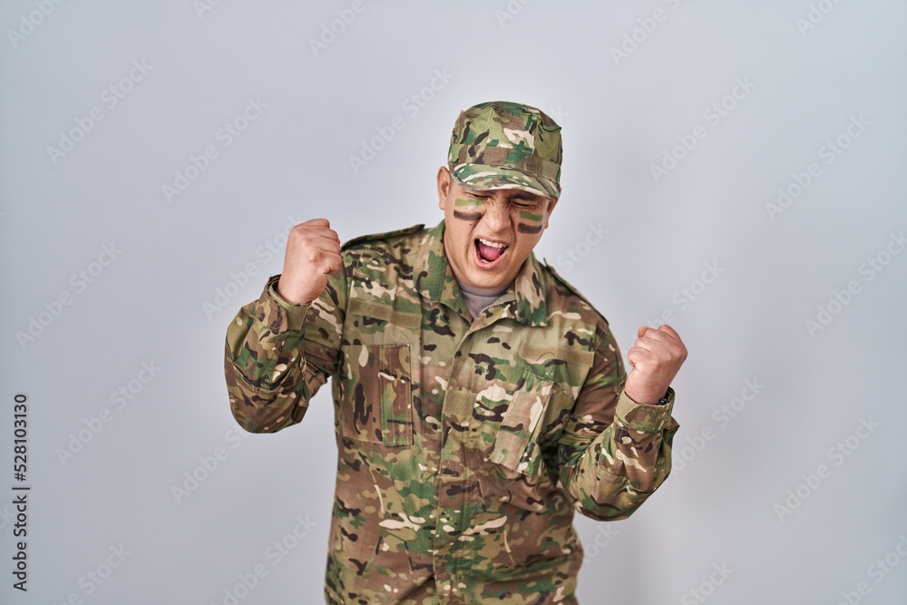 Hispanic young man wearing camouflage army uniform very happy and excited doing winner gesture with arms raised, smiling and screaming for success. celebration concept. Hispanic young man wearing camouflage army uniform very happy and excited doing winner gesture with arms raised, smiling and screaming for success. celebration concept.