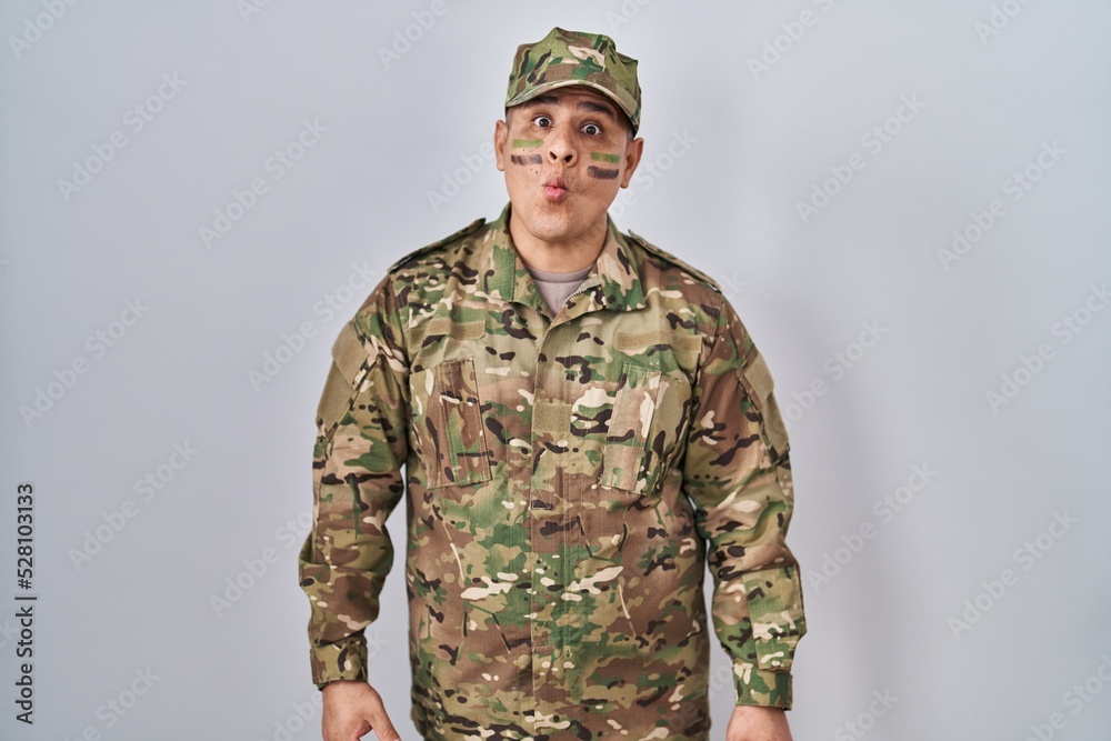 Hispanic young man wearing camouflage army uniform making fish face with lips, crazy and comical gesture. funny expression. Hispanic young man wearing camouflage army uniform making fish face with lips, crazy and comical gesture. funny expression.