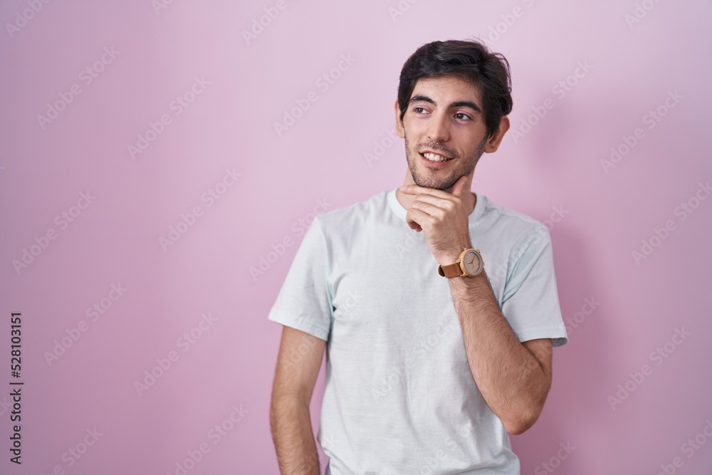 Young hispanic man standing over pink background with hand on chin thinking about question, pensive expression. smiling and thoughtful face. doubt concept. Young hispanic man standing over pink background with hand on chin thinking about question, pensive expression. smiling and thoughtful face. doubt concept.