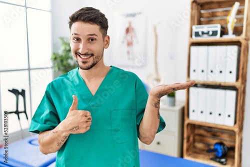 Young physiotherapist man working at pain recovery clinic showing palm hand and doing ok gesture with thumbs up, smiling happy and cheerful