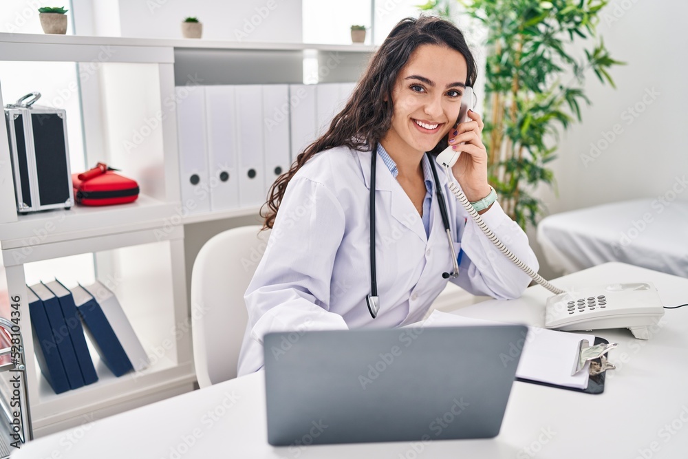 Young hispanic woman wearing doctor uniform talking on the telephone at clinic Young hispanic woman wearing doctor uniform talking on the telephone at clinic