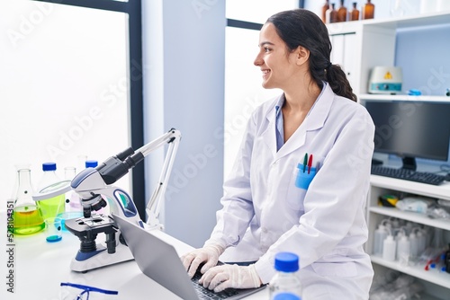 Young hispanic woman wearing scientist uniform using laptop at laboratory
