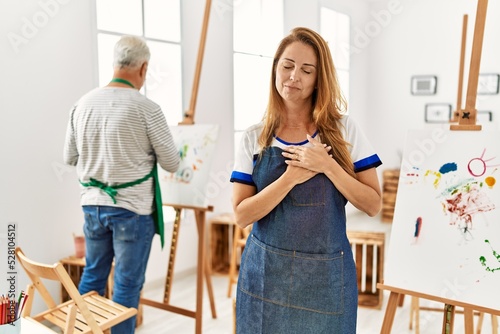 Hispanic woman wearing apron at art studio smiling with hands on chest with closed eyes and grateful gesture on face. health concept.