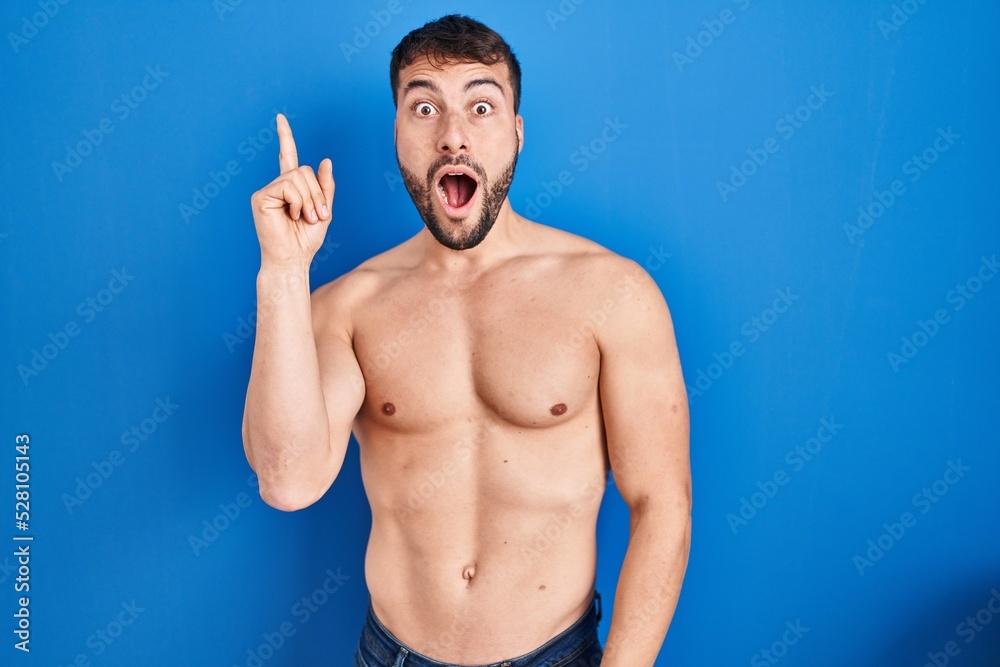Fototapeta premium Handsome hispanic man standing shirtless pointing finger up with successful idea. exited and happy. number one.
