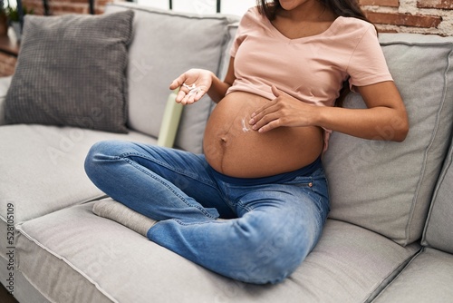 Young latin woman pregnant applying cream lotion on skin belly at home