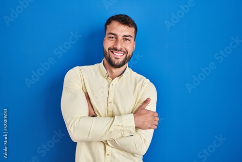 Handsome hispanic man standing over blue background happy face smiling with crossed arms looking at the camera. positive person.