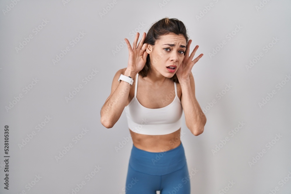 Hispanic woman wearing sportswear over isolated background trying to hear both hands on ear gesture, curious for gossip. hearing problem, deaf