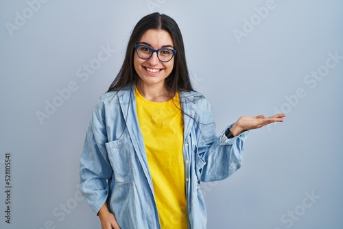 Young hispanic woman standing over blue background smiling cheerful presenting and pointing with palm of hand looking at the camera.