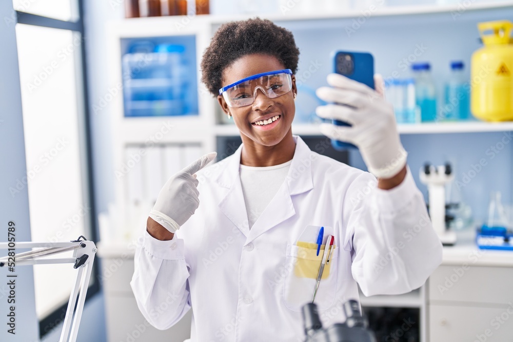 African american woman working at scientist laboratory doing video call with smartphone smiling happy pointing with hand and finger African american woman working at scientist laboratory doing video call with smartphone smiling happy pointing with hand and finger