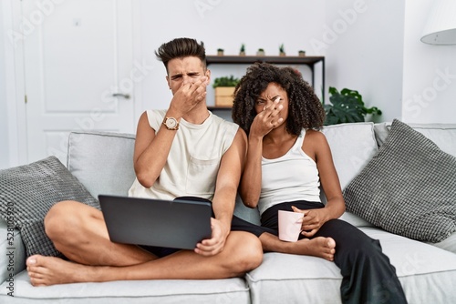 Young interracial couple using laptop at home sitting on the sofa smelling something stinky and disgusting, intolerable smell, holding breath with fingers on nose. bad smell