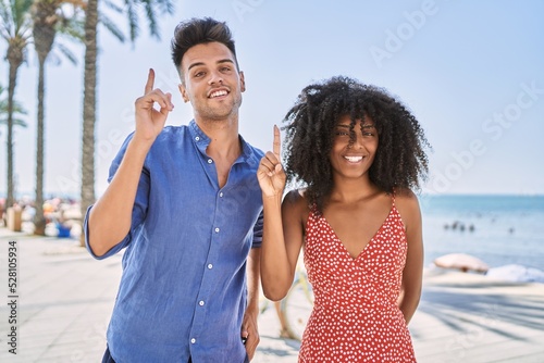Young interracial couple outdoors on a sunny day pointing finger up with successful idea. exited and happy. number one.