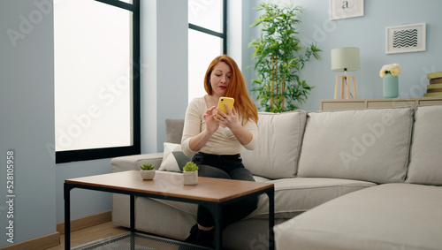 Young redhead woman using smartphone sitting on sofa at home