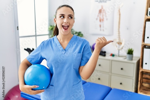 Young physiotherapist woman holding pilates ball at medical clinic pointing thumb up to the side smiling happy with open mouth