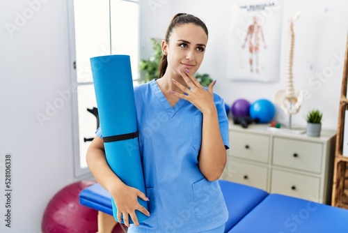 Young physiotherapist woman holding yoga mat serious face thinking about question with hand on chin  thoughtful about confusing idea