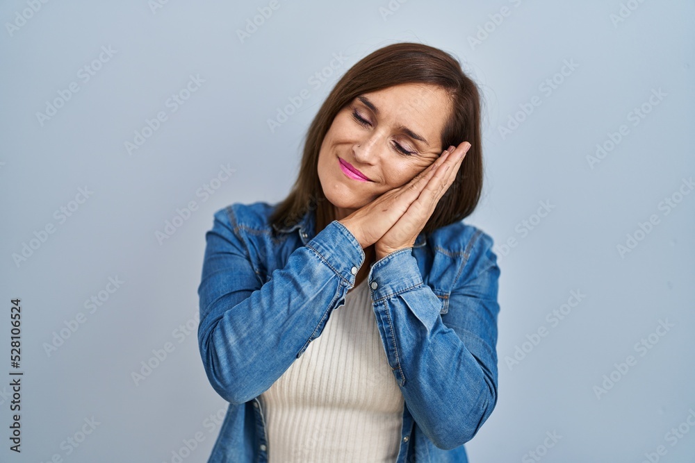 Middle age brunette woman wearing casual denim jacket over isolated background sleeping tired dreaming and posing with hands together while smiling with closed eyes. Middle age brunette woman wearing casual denim jacket over isolated background sleeping tired dreaming and posing with hands together while smiling with closed eyes.