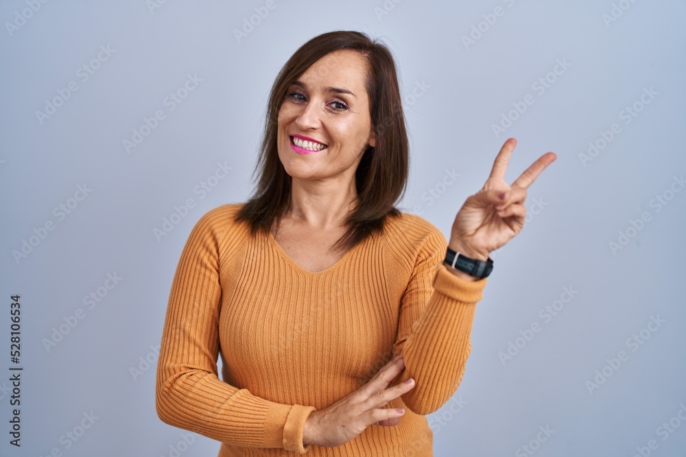 Middle age brunette woman standing wearing orange sweater smiling looking to the camera showing fingers doing victory sign. number two. Middle age brunette woman standing wearing orange sweater smiling looking to the camera showing fingers doing victory sign. number two.