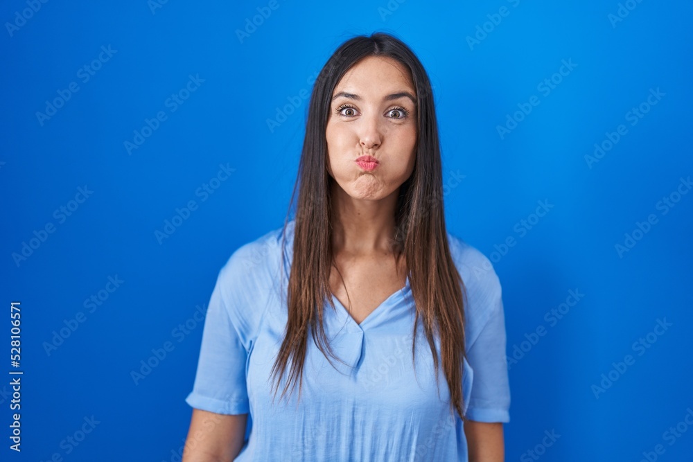 Young brunette woman standing over blue background puffing cheeks with funny face. mouth inflated with air, crazy expression. Young brunette woman standing over blue background puffing cheeks with funny face. mouth inflated with air, crazy expression.