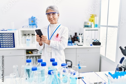 Middle age grey-haired woman wearing scientist uniform using smartphone at laboratory