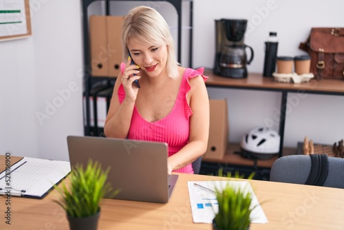 Young blonde woman business worker using laptop talking on smartphone at office