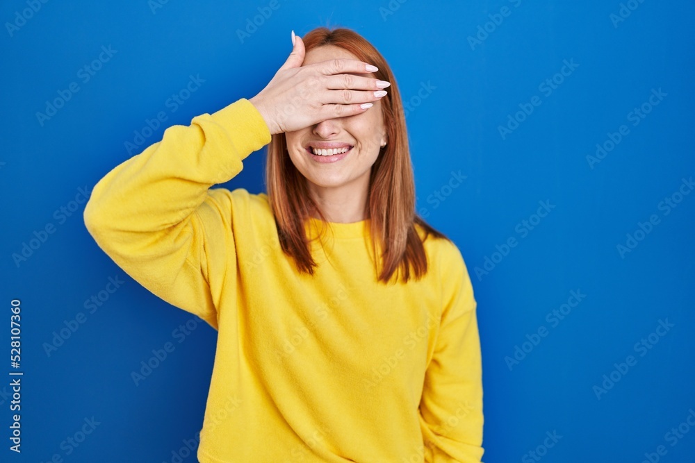 Young woman standing over blue background smiling and laughing with hand on face covering eyes for surprise. blind concept.