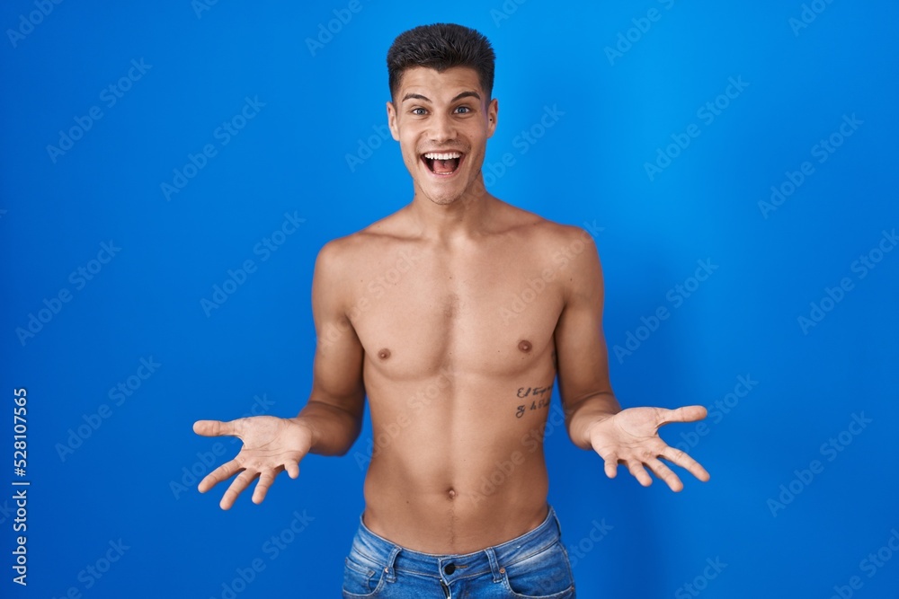 Young hispanic man standing shirtless over blue background smiling cheerful with open arms as friendly welcome, positive and confident greetings