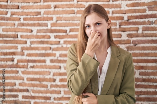 Young caucasian woman standing over bricks wall background smelling something stinky and disgusting, intolerable smell, holding breath with fingers on nose. bad smell