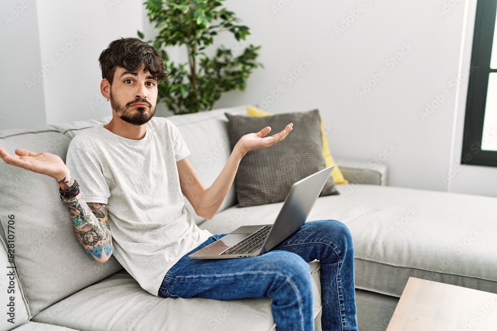 Hispanic man with beard sitting on the sofa clueless and confused expression with arms and hands raised. doubt concept.