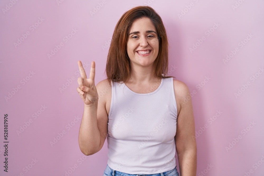 Brunette woman standing over pink background showing and pointing up with fingers number two while smiling confident and happy. Brunette woman standing over pink background showing and pointing up with fingers number two while smiling confident and happy.