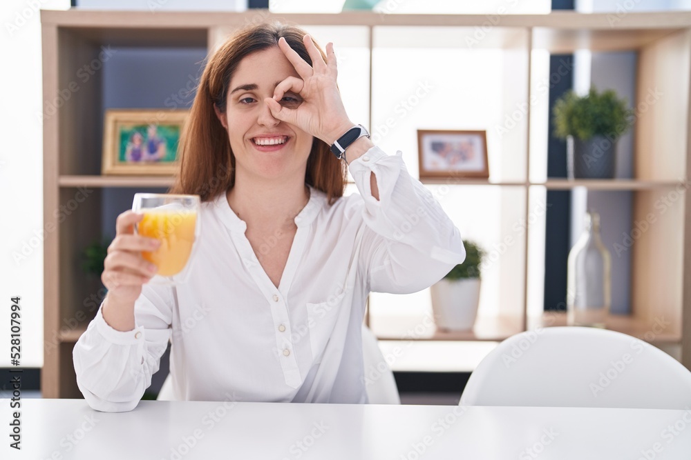 Brunette woman drinking glass of orange juice doing ok gesture with hand smiling, eye looking through fingers with happy face. Brunette woman drinking glass of orange juice doing ok gesture with hand smiling, eye looking through fingers with happy face.