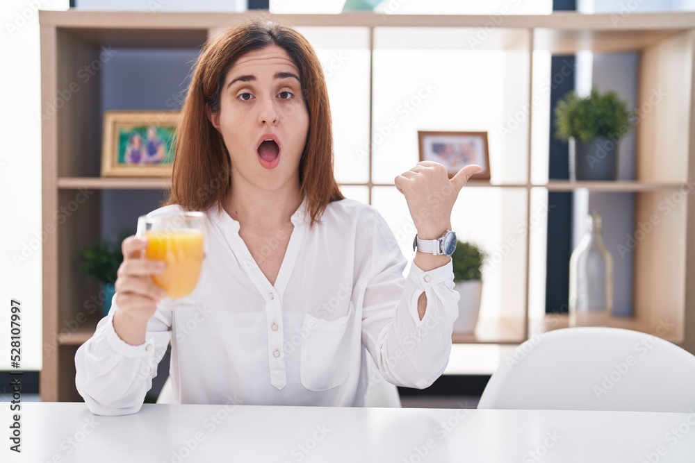 Brunette woman drinking glass of orange juice surprised pointing with hand finger to the side, open mouth amazed expression. Brunette woman drinking glass of orange juice surprised pointing with hand finger to the side, open mouth amazed expression.