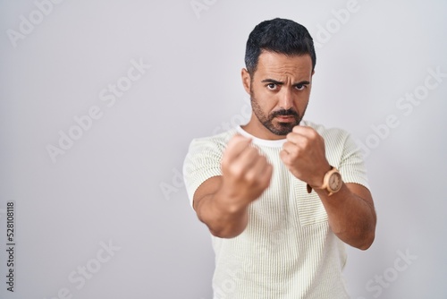 Hispanic man with beard standing over isolated background ready to fight with fist defense gesture, angry and upset face, afraid of problem