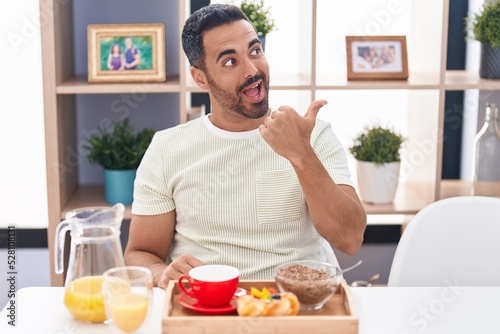 Hispanic man with beard eating breakfast pointing thumb up to the side smiling happy with open mouth