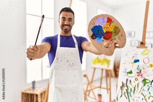 Young hispanic man smiling confident holding paintbrush and palette at art studio