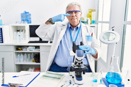 Senior caucasian man working at scientist laboratory pointing with hand finger to face and nose, smiling cheerful. beauty concept