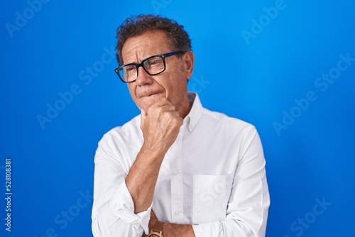 Middle age hispanic man standing over blue background looking confident at the camera smiling with crossed arms and hand raised on chin. thinking positive. © Krakenimages.com
