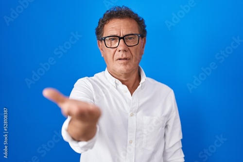 Middle age hispanic man standing over blue background smiling cheerful offering palm hand giving assistance and acceptance.