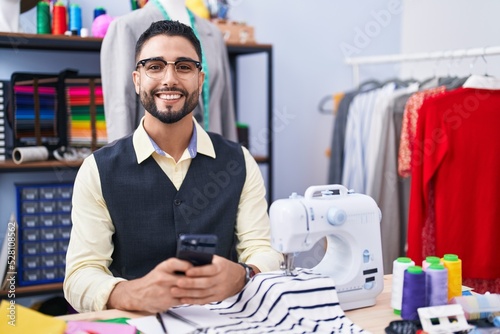Young arab man tailor smiling confident using smartphone at clothing factory