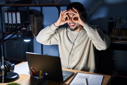 Young handsome man working using computer laptop at night doing ok gesture like binoculars sticking tongue out, eyes looking through fingers. crazy expression.