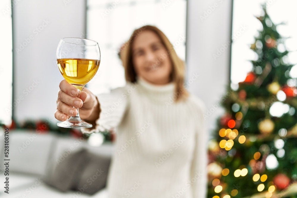 Middle age caucasian woman toasting with champagne standing by christmas tree at home