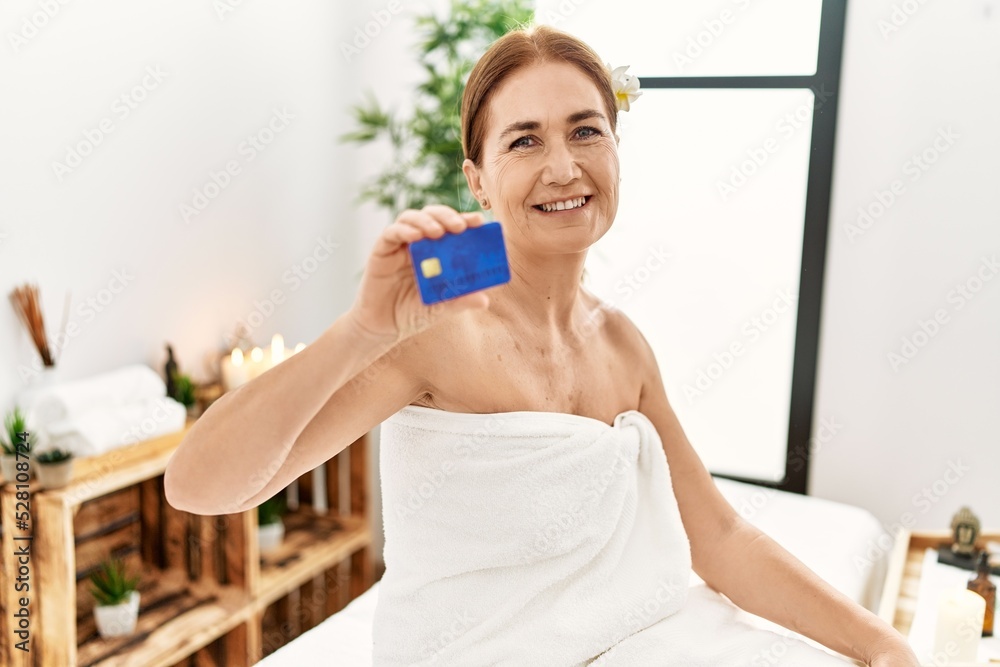 Middle age caucasian woman wearing towel holding credit card sitting on massage board at beauty center Middle age caucasian woman wearing towel holding credit card sitting on massage board at beauty center