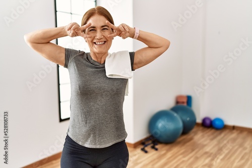 Middle age woman wearing sporty look training at the gym room doing peace symbol with fingers over face, smiling cheerful showing victory