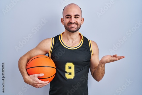 Young bald man with beard wearing basketball uniform holding ball smiling cheerful presenting and pointing with palm of hand looking at the camera.